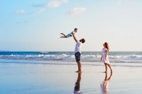 Happy family holidays. Joyful father, mother, baby son walk with fun along edge of sunset sea surf on black sand beach. Active parents and people outdoor activity on summer vacations with children.